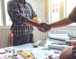 a contractor shaking another persons hand over a table. One is holding a hard had under his arm.