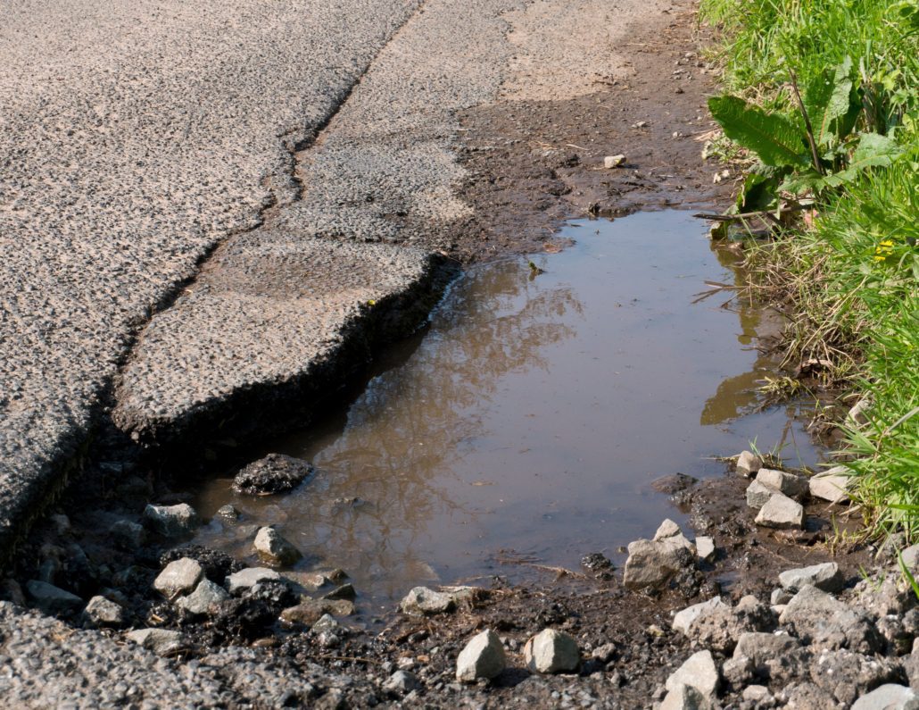 A pothole in the road by a grass verge with water in it.